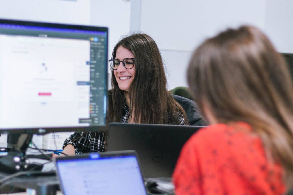 OpenSistemas - Consultora IT 205 Una escena de oficina moderna en la que dos mujeres trabajan frente a sus ordenadores portátiles. En primer plano, desenfocada, se ve una persona de espaldas con una blusa roja, al fondo, en el centro de la composición, destaca una mujer con gafas y cabello oscuro, largo y suelto que sonríe mientras mira la pantalla de su portátil.