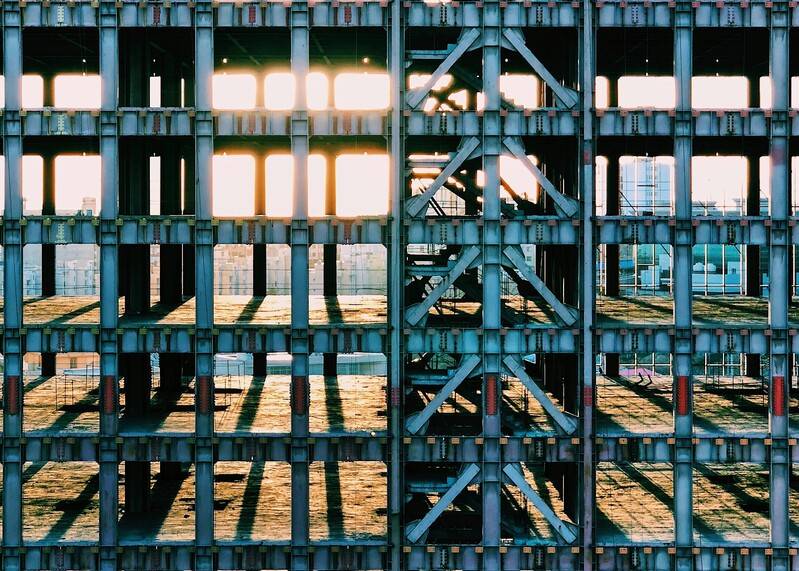 OpenSistemas - IT Consultancy 50 La estructura metálica de un edificio en construcción o quizá abandonado. Se observan vigas y columnas de acero organizadas en una retícula rectangular que deja ver el interior vacío. La luz del sol atraviesa los huecos, proyectando largas sombras sobre el suelo y creando un fuerte efecto de líneas y contrastes entre luz y sombra.