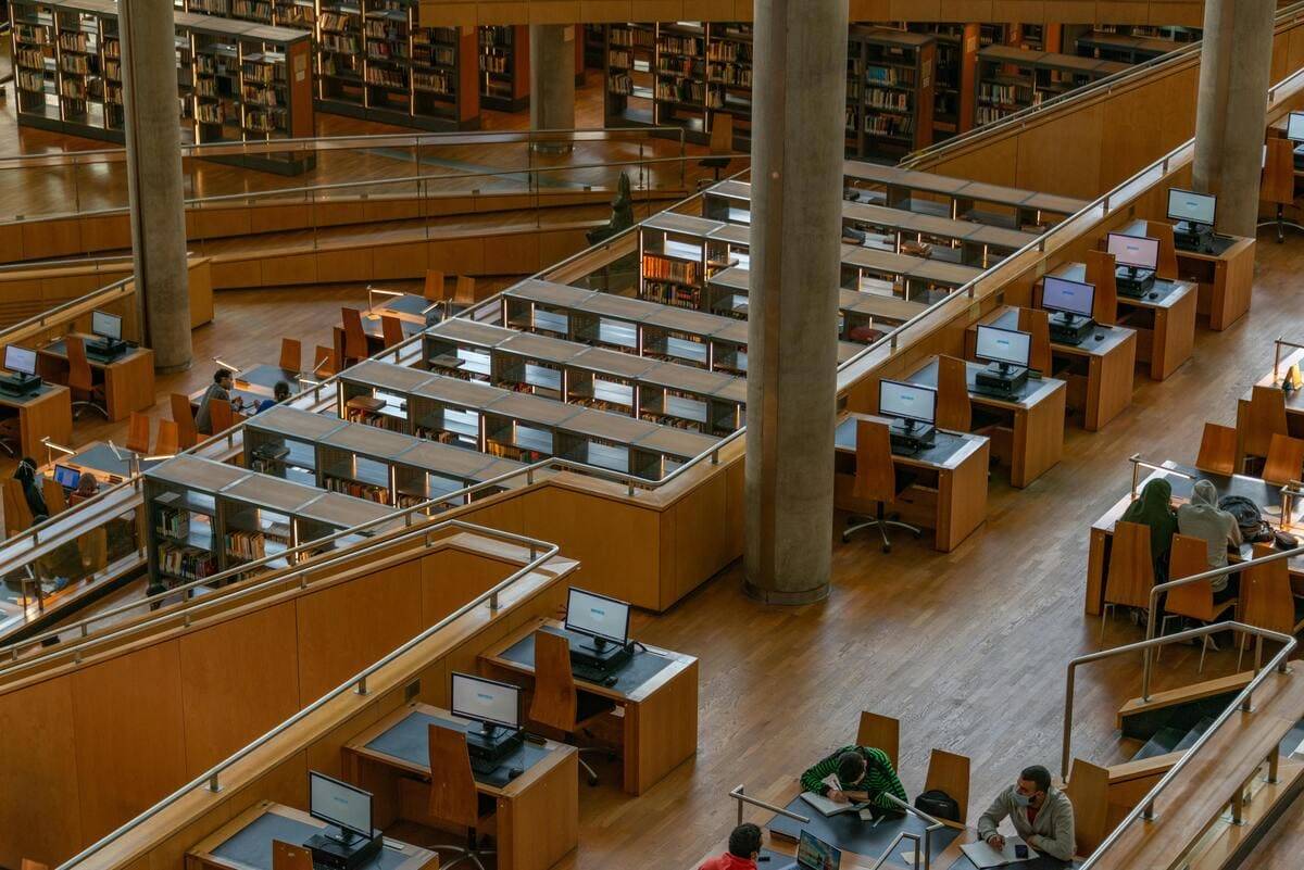 Una biblioteca amplia y moderna, el espacio está organizado en varios niveles con estanterías llenas de libros y áreas de trabajo individuales equipadas con computadoras de escritorio.