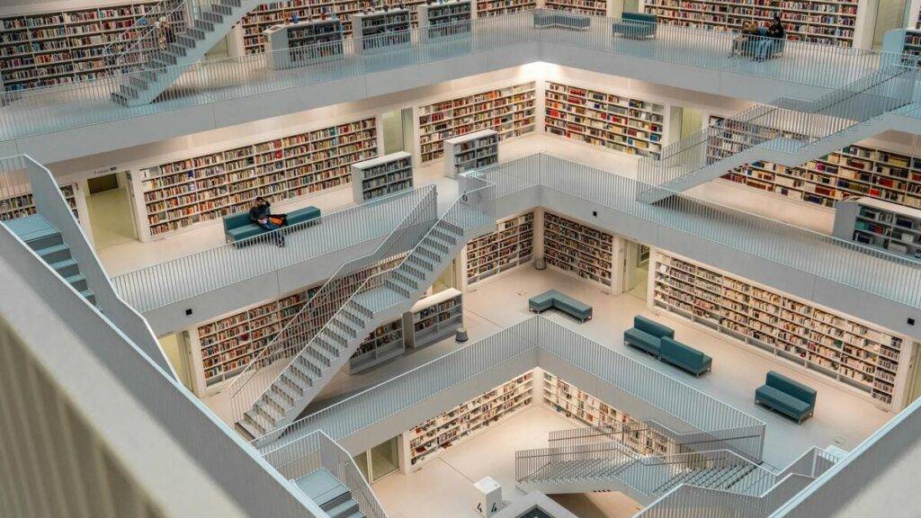 OpenSistemas - Consultora IT 68 Una biblioteca moderna y minimalista, caracterizada por su arquitectura abierta y geométrica. El espacio está organizado en varios niveles conectados por escaleras blancas, con estanterías repletas de libros que cubren las paredes de cada planta.